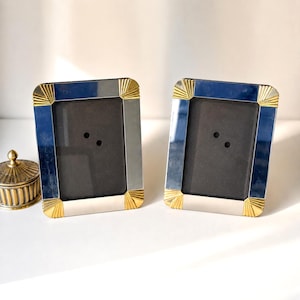 May include: Two rectangular picture frames with polished silver-tone sides and gold-tone sunburst corner accents, featuring dark gray inserts. Beside them sits a small, round, ribbed brass container with a domed lid and a pointed finial top.