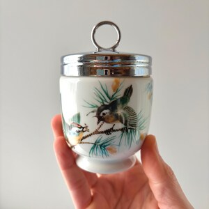 May include: A white porcelain egg coddler featuring a painted design of two small birds perched on pine branches with green needles. The container has a polished silver-tone metal lid with a circular ring handle on top.