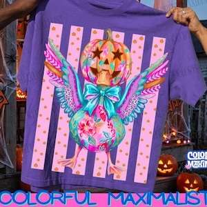 May include: A purple t-shirt featuring a graphic of a duck with a pumpkin head, star-shaped eyes, and a teal bow. The duck has colorful floral and leopard print wings. The background design consists of vertical pink stripes with small orange pumpkins.
