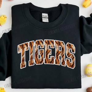 May include: Black sweatshirt with the word "TIGERS" in large, orange and brown tiger stripe lettering. The sweatshirt has a crew neck and long sleeves. The text "COLORFUL MAXIMALIST" is at the bottom of the image.