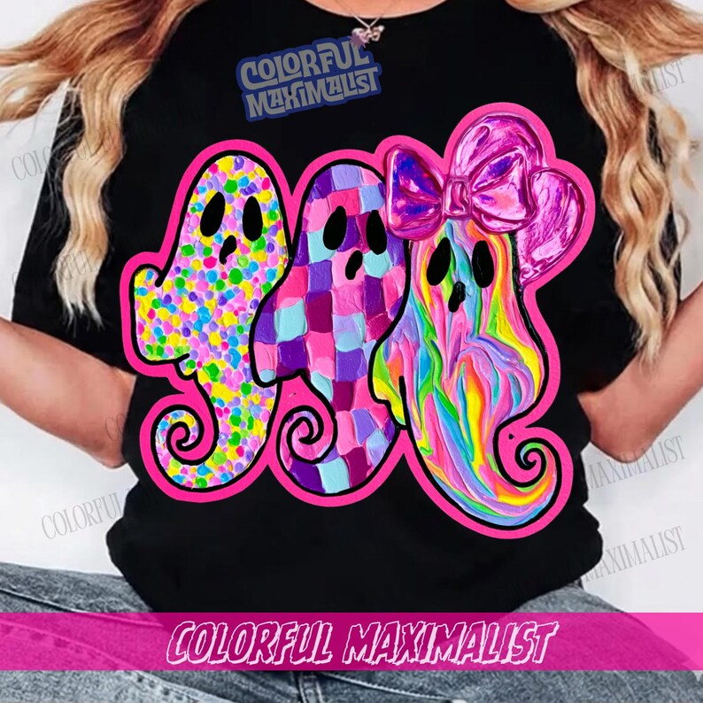 May include: A black t-shirt featuring a graphic of three ghosts outlined in bright pink. The first ghost has a multicolored dot pattern, the second a purple and blue checkered design, and the third a rainbow swirl pattern with a pink bow.
