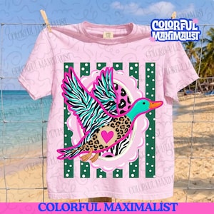 May include: A light pink t-shirt featuring a graphic of a flying duck with teal and black zebra stripes on its wings and a brown leopard print body. The duck has a bright pink heart on its chest and is centered over green vertical stripes with white dots.