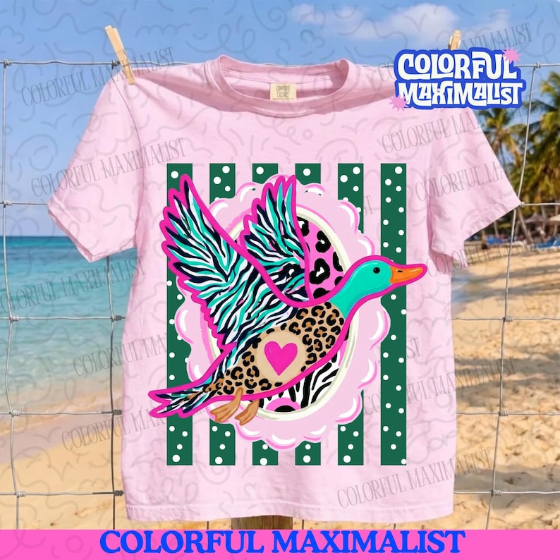 May include: A light pink t-shirt featuring a graphic of a flying duck with teal and black zebra stripes on its wings and a brown leopard print body. The duck has a bright pink heart on its chest and is centered over green vertical stripes with white dots.