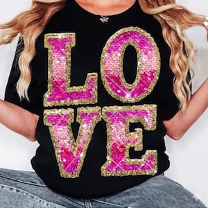 Puede incluir: Camiseta negra con la palabra "LOVE" escrita en letras grandes con lentejuelas. Las letras tienen un degradado de rosa y dorado, con un contorno dorado brillante. La camiseta es de estilo cuello redondo clásico.