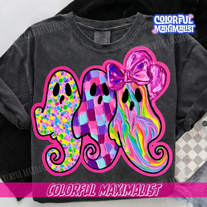 May include: A charcoal gray t-shirt featuring a graphic of three ghosts outlined in bright pink. The first ghost has a multicolored polka dot pattern, the second has a purple and blue checkered design, and the third has a pink bow and swirl pattern.