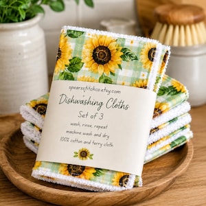 May include: A set of three dishwashing cloths featuring a yellow sunflower and green gingham pattern. The cloths are wrapped in a paper band labeled spearsstitchco.etsy.com, Dishwashing Cloths, Set of 3, wash, rinse, repeat, machine wash and dry.