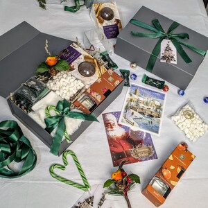 May include: A festive grey gift box tied with a green ribbon, containing Christmas goodies. The box includes chocolates, marshmallows, a candle, and a Christmas card. Candy canes and other gifts are also visible.