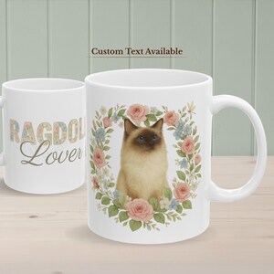 May include: White ceramic mug with a Ragdoll cat illustration framed by a floral wreath of pink roses and blue flowers. The mug has the text "Ragdoll Lover" on the side. The text "Custom Text Available" is also visible.
