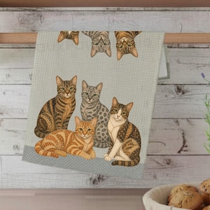 May include: A light green and white checked kitchen towel featuring a group of cats. The cats are depicted in various poses, with some upright and others upside down. The towel has a grey border at the bottom.