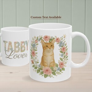 Puede incluir: Taza de cerámica blanca con una ilustración de gato atigrado rodeada de una corona floral de flores rosas y azules. La taza de la izquierda tiene las palabras "TABBY Lover" impresas. El texto "Custom Text Available" está encima de la taza.