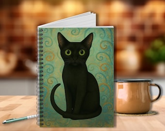 Op de afbeelding: Spiraalgebonden notitieboek met een illustratie van een zwarte kat op een turquoise achtergrond met gouden krullen. Een bruine mok en pen zijn ook zichtbaar op een houten oppervlak. Het notitieboek is open en toont blanco witte pagina's.