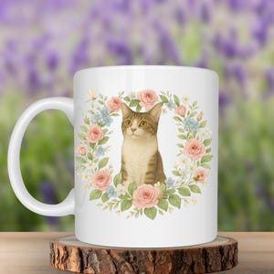Könnte beinhalten: Weiße Keramiktasse mit einer Aquarellillustration einer getigerten Katze, die von einem Blumenkranz aus rosa Rosen, blauen Vergissmeinnicht und Grün umgeben ist. Die Tasse hat einen gebogenen Henkel und steht auf einem Holzuntersetzer.