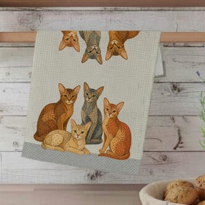 May include: A light blue and white checked kitchen towel with illustrations of cats in various poses. The cats are in shades of orange, tan, and grey, with detailed fur patterns. The towel is hanging on a wooden bar.