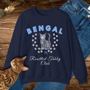 Könnte beinhalten: Marineblaues Sweatshirt mit dem Wort "BENGAL" in hellblauen, gebogenen Buchstaben. Darunter befindet sich eine Grafik einer Bengal-Katze, umgeben von weißen Pfotenabdrücken. Der Text "Rosetted Tabby Club" befindet sich unter der Katzen-Grafik.