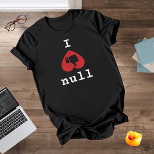 以下が含まれることがあります： 「I null」の白文字と赤いハートと親指を下向きにしたグラフィックが特徴の黒いTシャツ。木製の表面に置かれ、ノートパソコン、メガネ、アヒルのラバーダックが近くにあります。