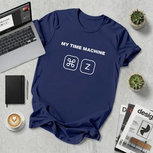 May include: A navy blue t-shirt featuring the text "MY TIME MACHINE" and the Command and Z keys in white. The shirt is on a light grey surface, with a laptop, notebook, pen, coffee, and magazines nearby.