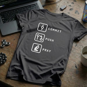 Peut inclure: T-shirt gris foncé avec texte et graphiques blancs. Le texte indique "COMMIT, PUSH, PRAY" avec des icônes correspondantes. Le t-shirt est posé sur une surface en bois avec un ordinateur portable, un fidget spinner et d'autres objets.