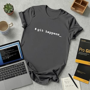 Peut inclure: T-shirt gris foncé avec le texte "$ git happens_" en blanc. Le t-shirt est présenté avec un ordinateur portable, des livres, une tasse et une petite plante succulente. C'est un vêtement décontracté.