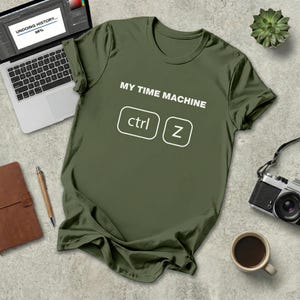 Puede incluir: Camiseta verde oliva con el texto "MY TIME MACHINE" y un gráfico "ctrl Z". También se ven un portátil, una cámara, una taza de café y una suculenta.