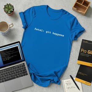Peut inclure: Un t-shirt bleu vif avec l'inscription blanche "fatal: git happens". L'image comprend un ordinateur portable, des livres, une tasse et une petite plante succulente. Le t-shirt est un vêtement décontracté.