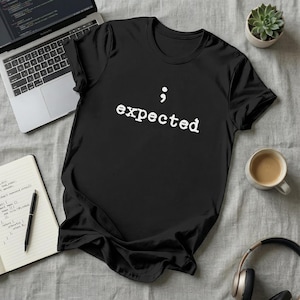 May include: Black t-shirt with the word "expected" in white, along with a semicolon symbol. The shirt is displayed on a gray surface, with a laptop, notebook, pen, coffee cup, headphones, and a small potted succulent nearby.