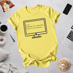 May include: A yellow t-shirt featuring a black computer monitor graphic with the text: "Error: Keyboard not connected. Press F1 to continue." The shirt has a crew neck and is made of a soft material.
