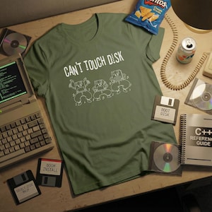 May include: An olive green t-shirt with the text "CAN'T TOUCH DISK" and a graphic of dancing computer characters. The image includes floppy disks, CDs, a vintage laptop, and a snack bag.