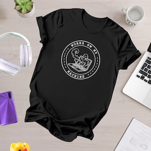 以下が含まれることがあります： 「WORKS ON MY MACHINE」の文字と、煙を出すノートパソコンの白いグラフィックがプリントされた黒いTシャツ。デザインはTシャツの中央に配置されています。Tシャツの襟が見えています。