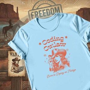 May include: Light blue t-shirt with a graphic of a cowgirl and the words "Coding Outlaw" in orange. The shirt also has the text "Born to Deploy on Fridays". The design has a vintage, western theme.