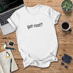 以下が含まれることがあります： 「got root?」の黒い文字が入った白いTシャツ。木製の表面に、ラップトップ、コーヒーカップ、多肉植物、その他のテクノロジー関連のアクセサリーと一緒に表示されています。