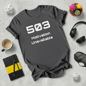 Könnte beinhalten: Dunkelgraues T-Shirt mit dem weißen Text "503 Motivation Unavailable". Das Bild zeigt auch eine Kaffeetasse, ein Notizbuch, einen Stift, Kopfhörer, eine kleine Topfpflanze und eine Schlafmaske.
