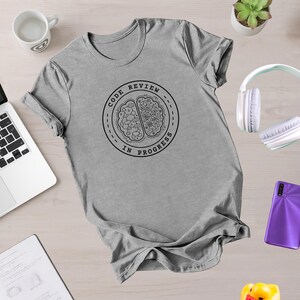 May include: A heather gray t-shirt with a round graphic that says "CODE REVIEW IN PROGRESS" around a brain illustration. The shirt is laid flat on a light wood-grain surface, with a laptop, mug, and headphones nearby.