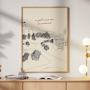 May include: A framed art print with a sketch of a town landscape, with the text "meet me in lawrence". The print hangs above a light wood cabinet. A gold lamp with spherical bulbs and a gold sculpture are on the cabinet.