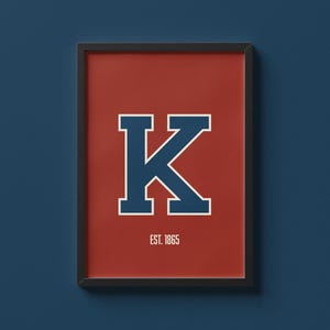 May include: Framed print featuring a large, navy blue letter "K" with a white outline on a red background. The text "EST. 1865" is below the letter. The print is in a black frame and hangs against a dark blue wall.