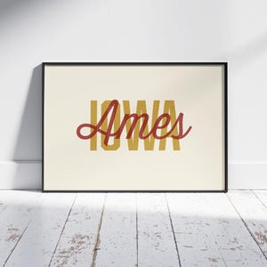 May include: A framed print featuring the word "IOWA" in a mustard yellow block font, with the word "Ames" in a cursive red font overlaid. The print is set against a cream background and framed in black, displayed on a white wall.
