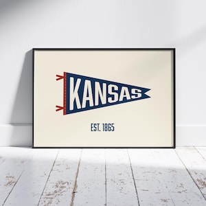 May include: A framed rectangular print featuring a dark blue triangular pennant with the word KANSAS in white capital letters. The pennant has red ties on the left side and the text EST. 1865 in dark blue centred below the pennant on a cream background.