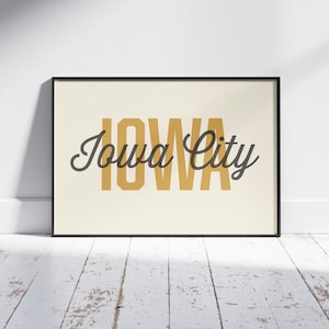 May include: A framed print with the words "IOWA" in a mustard yellow block font, with "Iowa City" in a cursive font overlaying the word "IOWA". The print is in a black frame and leans against a white wall.