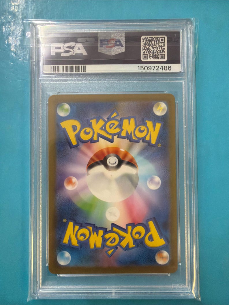 May include: A graded Pok&eacute;mon trading card encased in a clear plastic holder. The card's back features the Pok&eacute;mon logo in yellow and blue, with a white and red Pok&eacute; Ball design. The holder has a PSA label with a barcode and serial number.