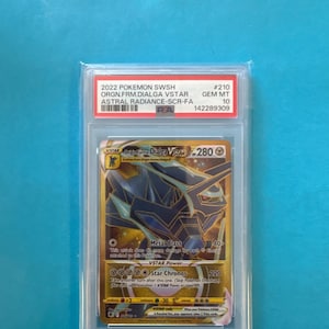 May include: A graded Pokémon card featuring Dialga VSTAR from the Astral Radiance set. The card has a gold and blue design with the text "Metal Blast" and "Star Chronos". The card is graded GEM MT 10 by PSA.