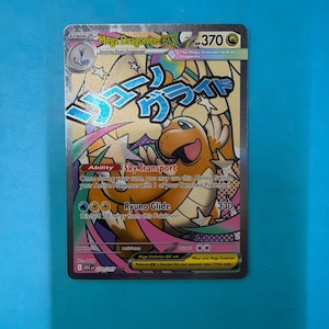 May include: A Mega Dragonite EX trading card with 370 HP, featuring a tan dragon-like creature, blue Japanese text, and a background of stars and colorful ribbons. The card displays the ability Sky Transport and the attack Ryuno Glide.