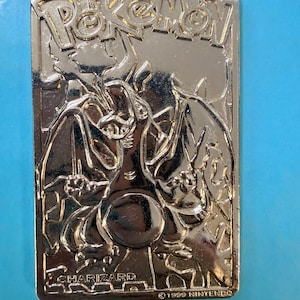 Puede incluir: Una tarjeta rectangular de metal color oro con una imagen en relieve del Pokémon Charizard. La parte superior muestra el logotipo de Pokémon, mientras que la inferior incluye el texto CHARIZARD y 1999 NINTENDO en relieve.