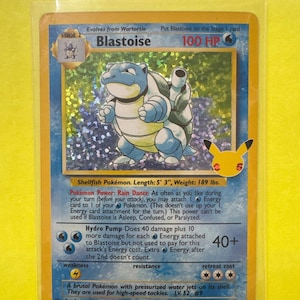 Può includere: Una carta Pokémon Blastoise con sfondo olografico. La carta presenta un personaggio Blastoise blu con un guscio e due cannoni ad acqua. Il testo della carta include gli HP, le abilità e i dettagli di attacco del personaggio. La carta è in una custodia protettiva.