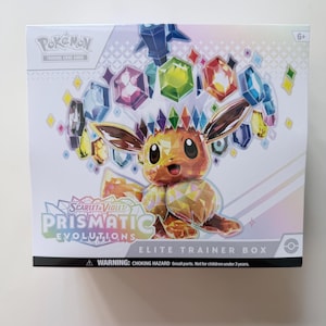 May include: A white Pokemon trading card game box featuring a colorful illustration of Eevee and gemstones. The box is labeled "Scarlet & Violet Prismatic Evolutions Elite Trainer Box" with a warning about small parts. The box is for ages 6+.