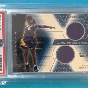 以下が含まれることがあります： 2001 SPx Winning Materialsシリーズの、グレーディングされたコービー・ブライアントのバスケットボールカード。レイカーズのユニフォームを着たブライアントの写真と、ジャージのスウォッチが特徴です。カードは保護ホルダーに収められています。
