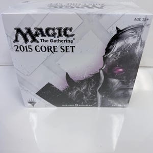 Puede incluir: Una caja Magic The Gathering 2015 Core Set Fat Pack con embalaje blanco, el logotipo del juego, texto que indica que incluye 9 sobres y un gráfico de un guerrero con casco oscuro y un ojo morado brillante en el lado derecho.