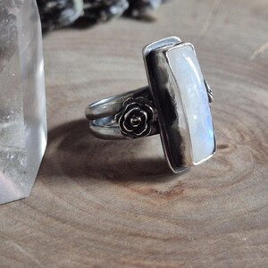 Puede incluir: Un anillo de plata con una piedra de luna rectangular y un dise&ntilde;o floral. El anillo est&aacute; sobre una superficie de madera.