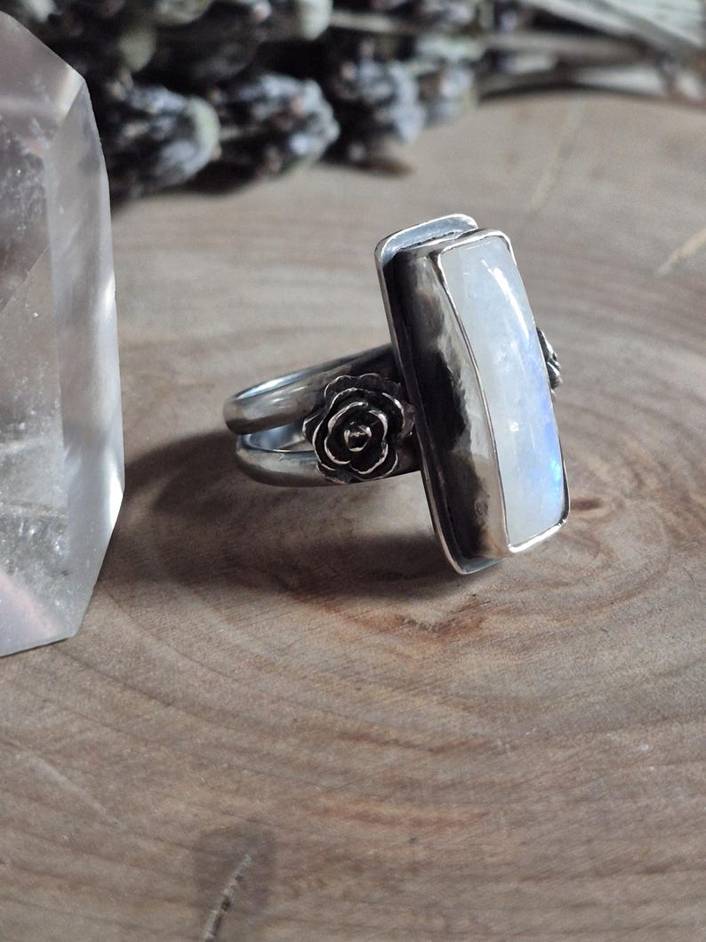 Puede incluir: Un anillo de plata con una piedra de luna rectangular y un dise&ntilde;o floral. El anillo est&aacute; sobre una superficie de madera.