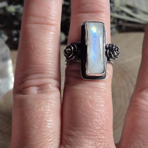 Puede incluir: Un anillo de plata con una piedra de luna rectangular engastada en un engaste de bisel. El anillo tiene dos peque&ntilde;os acentos en forma de rosa a cada lado de la piedra.