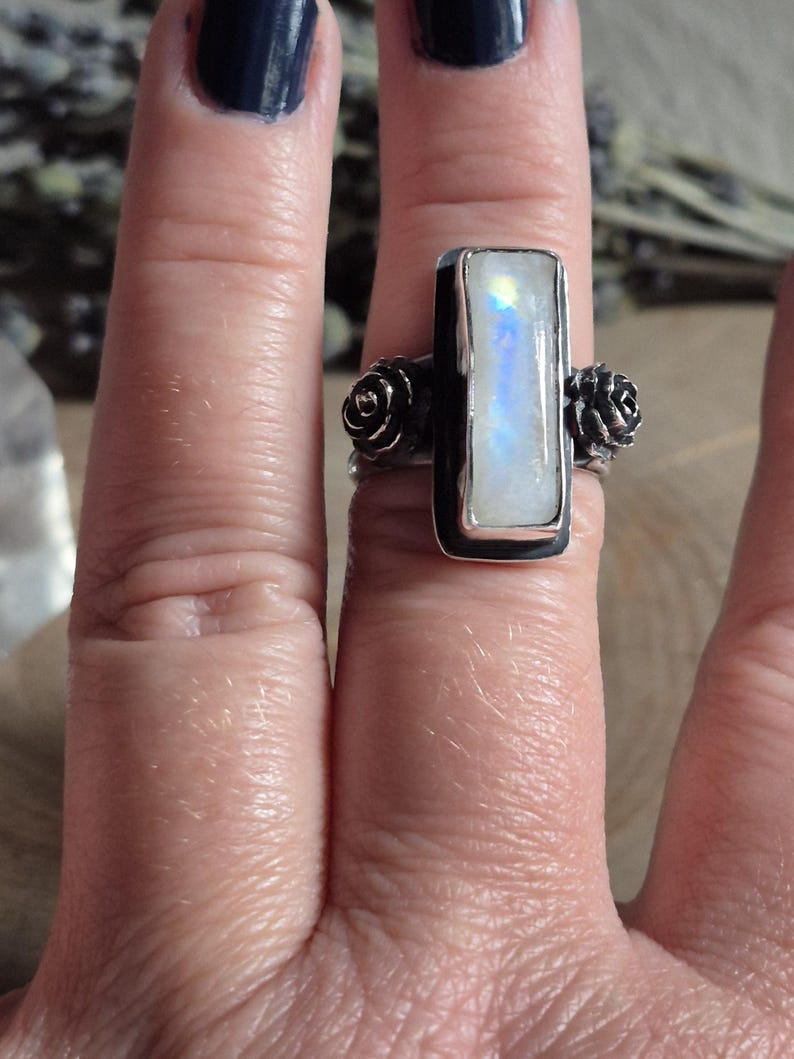 Puede incluir: Un anillo de plata con una piedra de luna rectangular engastada en un engaste de bisel. El anillo tiene dos peque&ntilde;os acentos en forma de rosa a cada lado de la piedra.