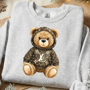 May include: A light gray sweatshirt featuring a watercolor illustration of a teddy bear wearing a brown hoodie with a repeating white logo pattern. The bear has a black nose and is sitting upright.